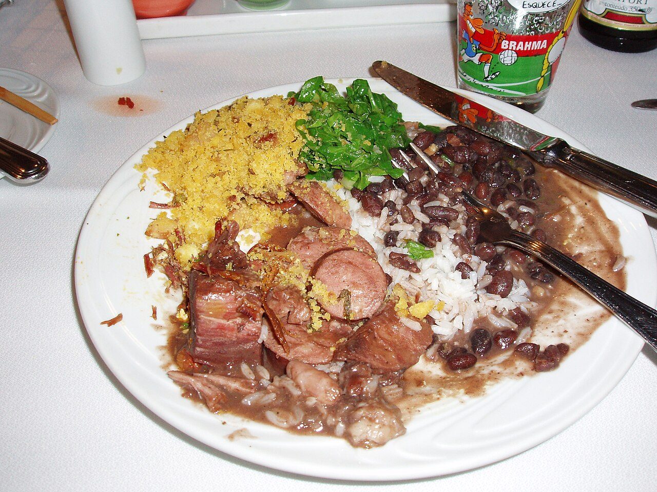 Prato branco de feijoada com arroz, feijão-preto, linguiça e carnes de porco, farofa e couve refogada, com um copo de cerveja ao fundo